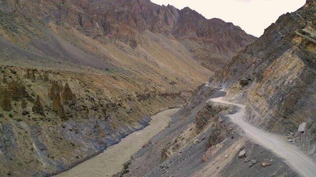 Aerial view of the winding Zanskar river flowing through a deep rocky canyon with a dirt road carved into the steep mountainside in Zanskar, Ladakh, India.