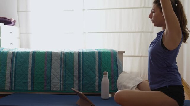 Athletic young woman performing stretching exercises while following an online yoga class on her tablet at home. After the workout, she drinks water