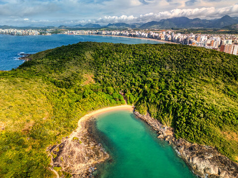 Aerial view of Praia do Ermitao featuring a secluded sandy beach nestled within lush green hills and rocky cliffs, with the coastal city skyline in the distance, Guarapari, Espirito Santo, Brazil.