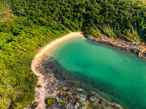 Aerial view of Praia do Ermitao featuring a secluded sandy beach with turquoise waters surrounded by lush green Atlantic forest and rocky coastline in Guarapari, Espirito Santo, Brazil.
