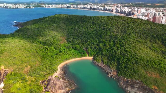Aerial view of Praia do Ermitao surrounded by lush forest with the city skyline of Guarapari in the distance, Guarapari, Espirito Santo, Brazil.