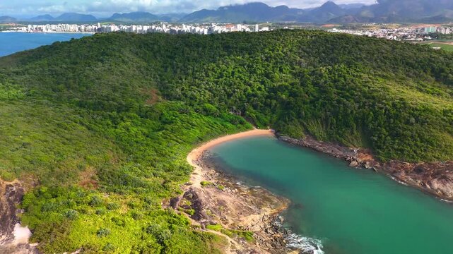 Aerial view of Praia do Ermitao with its turquoise cove and sandy beach surrounded by lush green hills and the city skyline of Guarapari, Espirito Santo, Brazil.