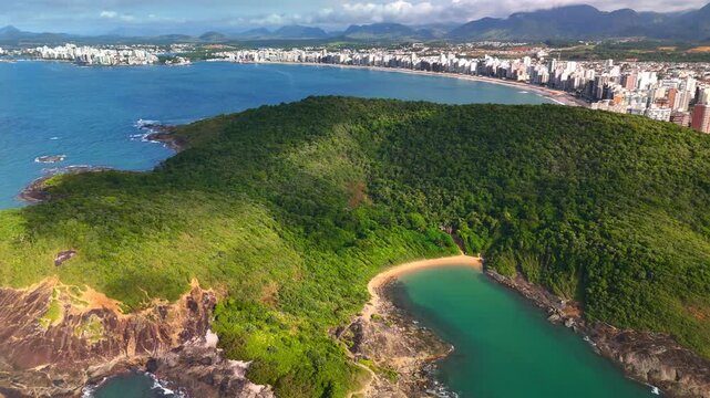 Aerial view of the lush green hills surrounding the secluded Praia do Ermitao beach with turquoise waters and the coastal skyline of Guarapari, Espirito Santo, Brazil.