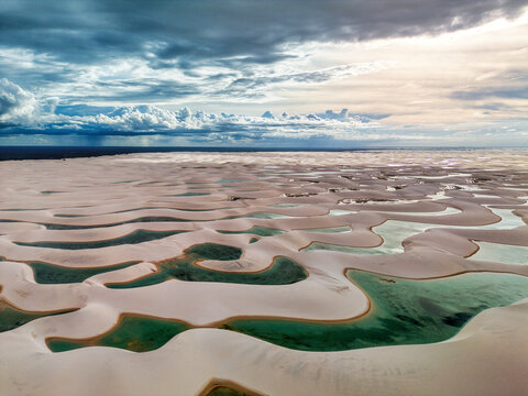 Aerial view of Lagoa Bonita featuring white sand dunes and turquoise rainwater lagoons under a dramatic cloudy sky in Barreirinhas, State of Maranhao, Brazil.