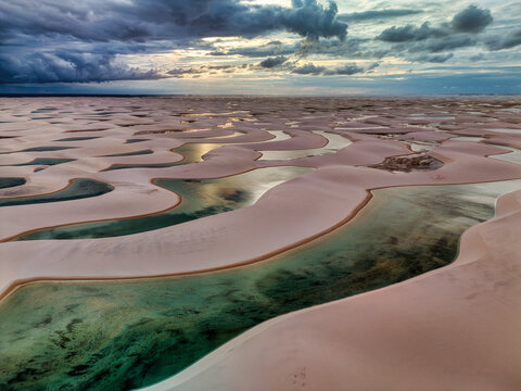 Aerial view of Lagoa Bonita with its crystal clear rainwater pools nestled among vast white sand dunes under a dramatic cloudy sky in Barreirinhas, Maranhao, Brazil.