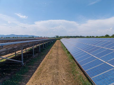 Aerial view of rows of photovoltaic solar panels installed in a field with a grassy path between them under a clear blue sky in Borgo Revel, Piedmont, Italy.