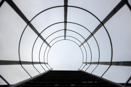 View of a metal ladder with a circular safety cage leading upwards against a bright overcast sky in Vilnius, Vilnius County, Lithuania.