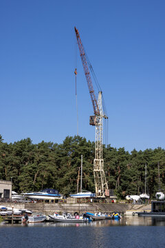 View of a tall construction crane standing by a waterfront with various boats docked, set against a backdrop of dense green trees under a clear blue sky in Vilnius, Vilnius County, Lithuania.
