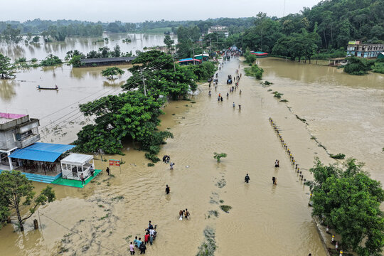 Satkania, Bangladesh - 11 July 2026: Aerial view of a flooded road in Satkania with people wading through brown floodwaters, surrounded by submerged trees and buildings.