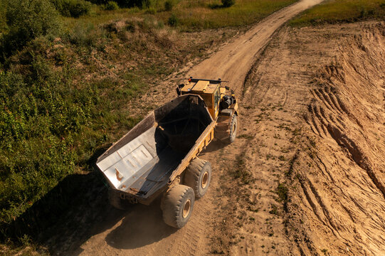 Aerial view of a large yellow dump truck driving along a dirt path in a quarry with sandy slopes and surrounding greenery in Naujoji Akmene, Siauliai County, Lithuania.