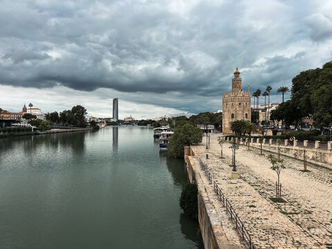Seville, Spain - 01 November 2025: View of the historic Torre del Oro standing along the Guadalquivir River in Seville, with a stone promenade, lush trees, and a cloudy sky.