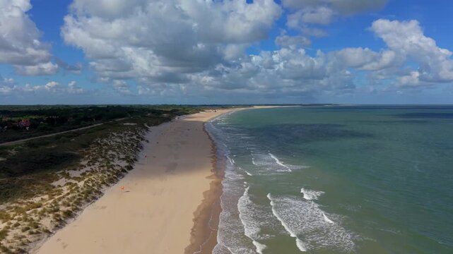 Aerial view video clip of expansive golden sandy beach, coastal sand dunes, North Sea ocean waves, and blue sky with cumulus clouds in Domburg, Zeeland, Netherlands on a sunny day