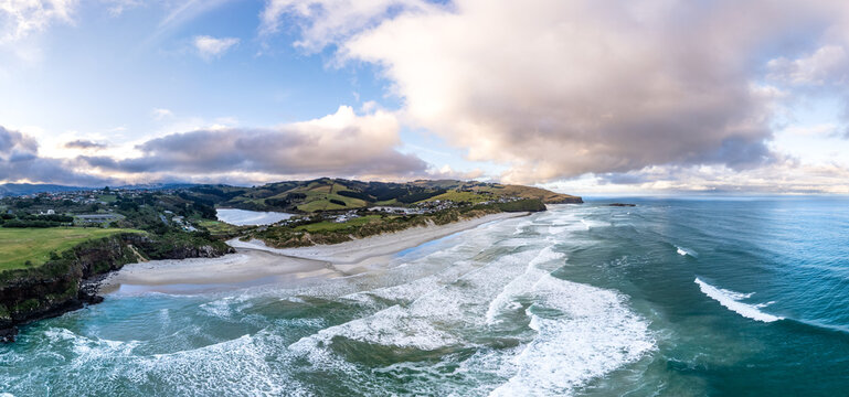 Aerial view of the coastline near Dunedin featuring sandy beaches, rolling green hills, and crashing ocean waves under a cloudy sky in Dunedin, Otago Region, New Zealand.