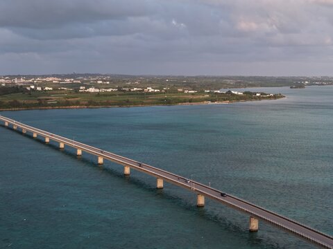 Aerial view of Irabu Bridge spanning the turquoise waters of the East China Sea with coastal greenery and cloudy sky in Miyakojima, Okinawa, Japan.