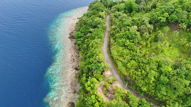 Aerial view of the coastal road winding through lush tropical forest near Lubang Buaya Morella with turquoise coral reef waters in Leihitu, Maluku, Indonesia.
