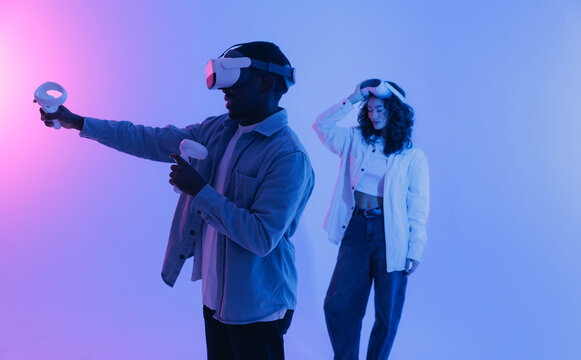 Young African male and Asian female engaging in virtual reality gaming with headsets and controllers, neon light
