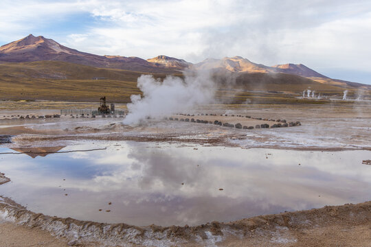 View of El Tatio geysers with steam rising from geothermal pools, volcanic mountains in the background, and reflections in the water in Antofagasta, Chile.