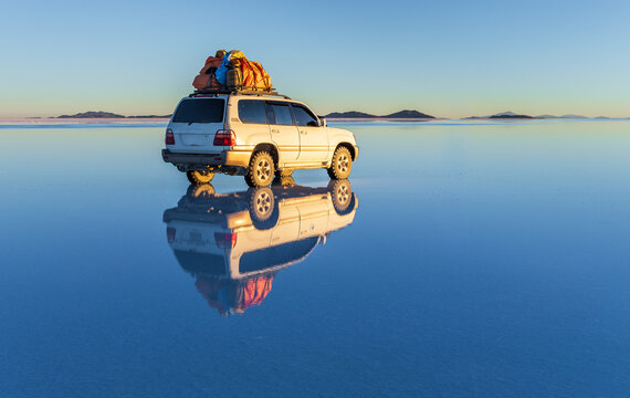 View of a white SUV with luggage on its roof reflected in the water of the Uyuni Salt Flat under a clear blue sky in Uyuni, Potosi Department, Bolivia.