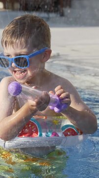 Little boy plays with colorful water blaster near swimming pool creating playful water spray. Water toy play, outdoor recreation, summer vacation and joyful childhood concept. 