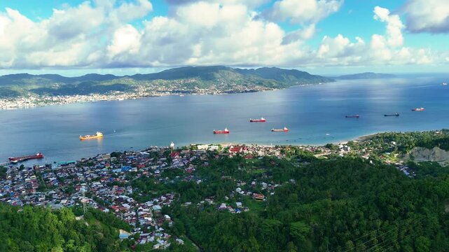 Aerial view of Teluk Ambon bay with cargo ships, coastal town, and lush green hills under a blue sky in Teluk Ambon, Maluku, Indonesia.
