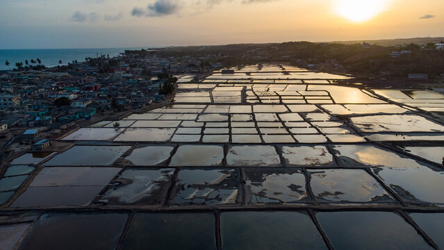 Aerial view of the salt pans in Elmina, reflecting the golden sunset light with the coastal town and ocean in the background, Elmina, Central Region, Ghana.
