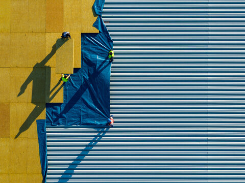 Aerial view of construction workers installing yellow insulation panels and blue protective sheeting on a corrugated metal roof in Belgrade, City of Belgrade, Serbia.