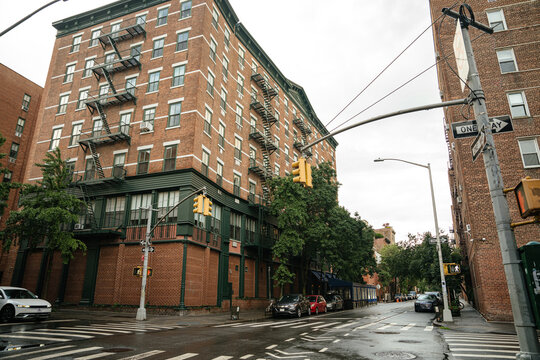 A classic New York City intersection featuring a red brick apartment building with metal fire escapes and traffic lights. Wet streets, crosswalk, and parked cars capture an authentic urban city