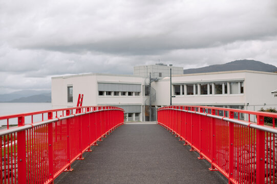 Modern white building with a bright red bridge leading towards it under cloudy skies