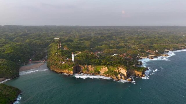 Aerial view of Baron Beach featuring the lighthouse perched on the cliffside above crashing waves and lush forest in Tanjungsari, Special Region of Yogyakarta, Indonesia.