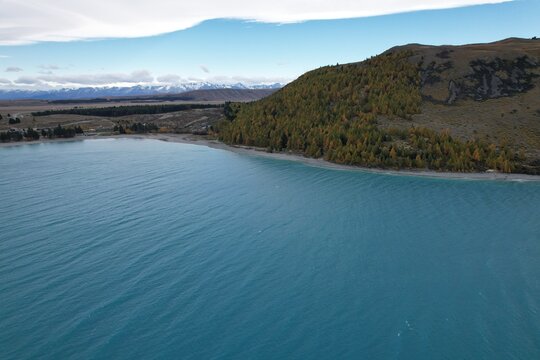 Aerial view of the turquoise waters of a lake bordered by a forested shoreline and rolling hills, with snow-capped mountains in the distance in Queenstown, Otago Region, New Zealand.