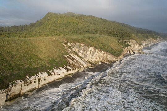 Aerial view of the rugged coastline of the South Island with dramatic layered cliffs, dark sandy beach, and crashing ocean waves in Queenstown, Otago Region, New Zealand.