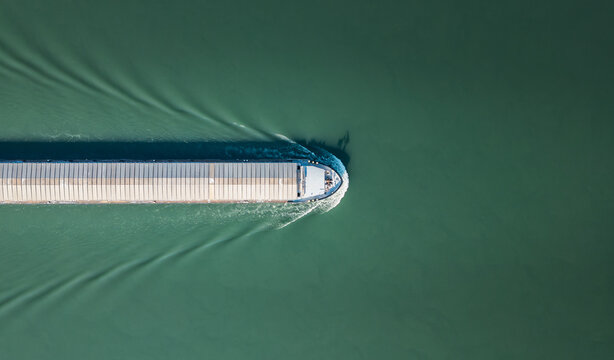 Aerial view of a cargo barge sailing on the Rhone River, creating symmetrical wake patterns and a distinct shadow on the water surface near Le Pouzin, Auvergne-Rhone-Alpes, France.