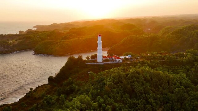 Aerial view of the white Baron Beach lighthouse perched on a cliff overlooking the ocean, surrounded by lush green hills during a golden sunset in Tanjungsari, Special Region of Yogyakarta, Indonesia.