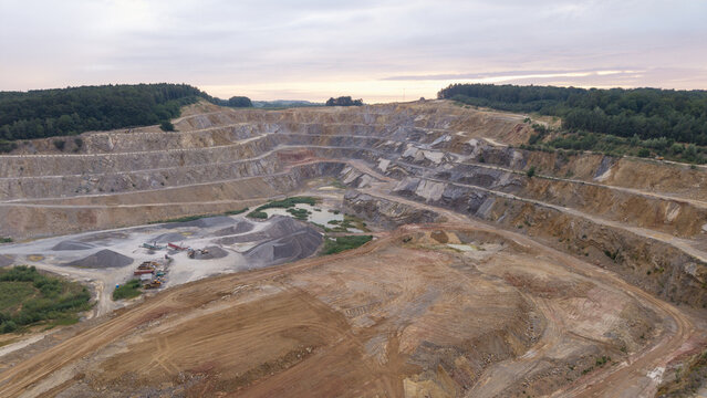 Aerial view of a large open-pit quarry with terraced rock walls, gravel piles, and a small pond surrounded by forest in the evening light in Krakow, Lesser Poland Voivode, Poland.