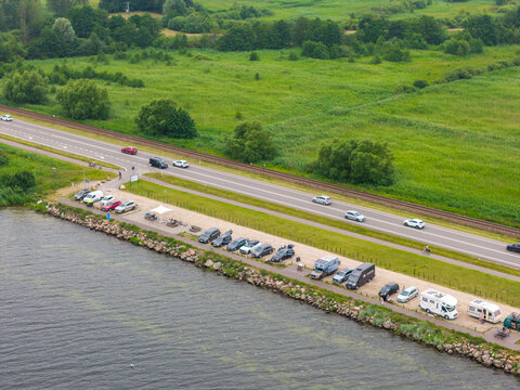 Aerial view of Puck Bay with a coastal parking area filled with cars and campers, adjacent to a road and railway line, surrounded by lush green fields in Puck, Pomeranian Voivodeship, Poland.