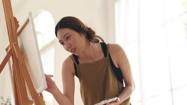 Creative young Asian woman artist painting on canvas in bright home studio. Relaxed female hobbyist enjoying peaceful indoor leisure time, expressing imagination and passion for art.