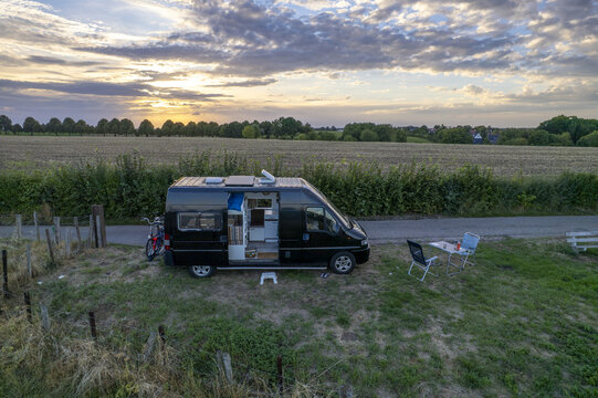 Aerial view of a black camper van parked in a grassy field with camping chairs and a table at sunset in Valkenburg, Limburg, Netherlands.