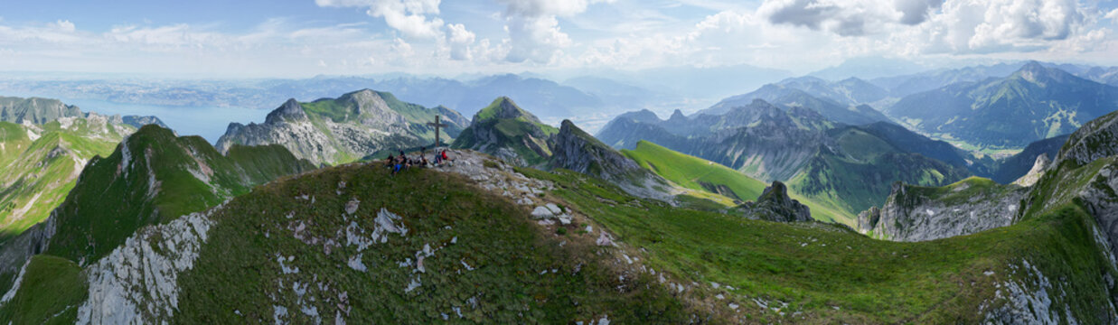Aerial view of Cornettes de Bise peak with a summit cross and hikers resting on the rocky ridge surrounded by green slopes in La Chapelle-d'Abondance, Auvergne-Rhone-Alpes, France.
