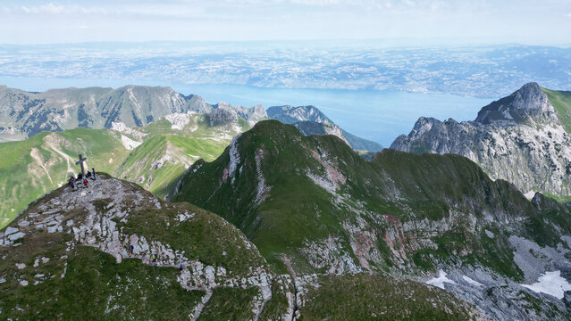 Aerial view of Cornettes de Bise peak with hikers near the summit cross, overlooking green mountain ridges and Lake Geneva in La Chapelle-d'Abondance, Auvergne-Rhone-Alpes, France.