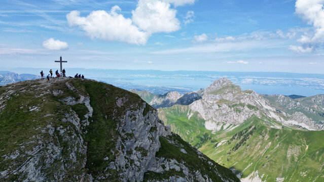 Aerial view of Cornettes de Bise summit with a cross and hikers on the rocky peak overlooking green slopes and a distant lake in La Chapelle-d'Abondance, Auvergne-Rhone-Alpes, France.
