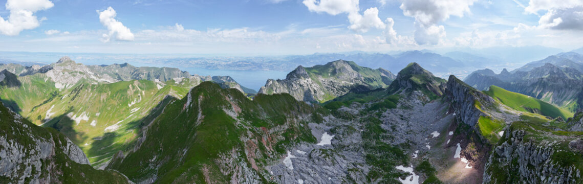 Aerial view of Cornettes de Bise mountain range with rugged rocky peaks, patches of snow, lush green alpine meadows, and distant blue lake in La Chapelle-d'Abondance, Auvergne-Rhone-Alpes, France.