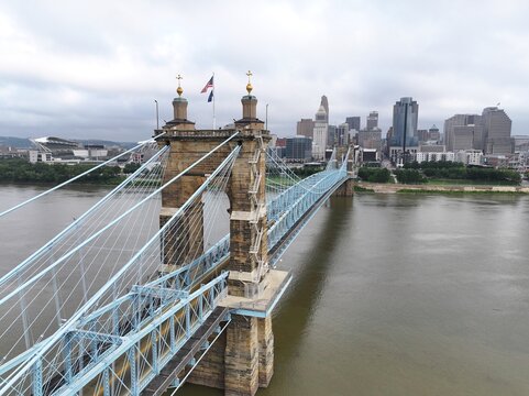 Cincinnati, United States - 12 July 2026: Aerial view of the John A. Roebling Suspension Bridge spanning the Ohio River with the Cincinnati skyline in the background under a cloudy sky.
