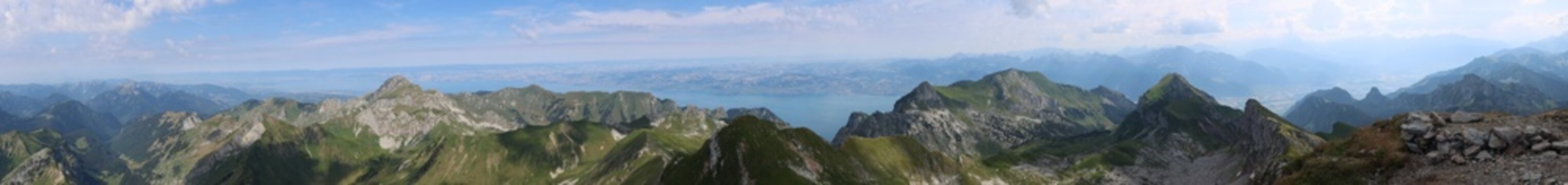 View of the Cornettes de Bise mountain range with rocky peaks, green slopes, and a distant lake under a blue sky in La Chapelle-d'Abondance, Auvergne-Rhone-Alpes, France.