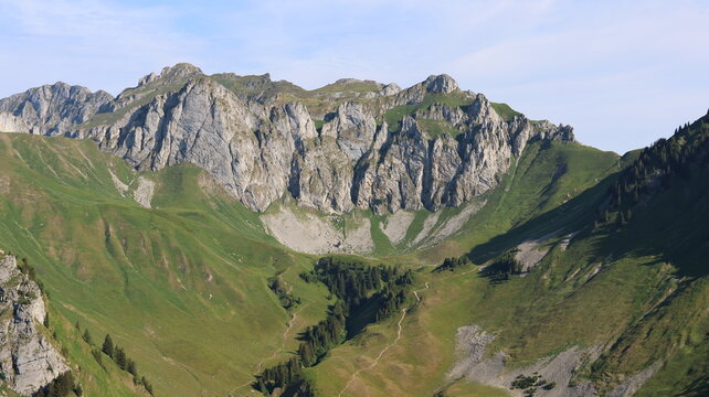 View of the Cornettes de Bise mountain range with lush green alpine meadows, rocky limestone peaks, and winding hiking trails in La Chapelle-d'Abondance, Auvergne-Rhone-Alpes, France.