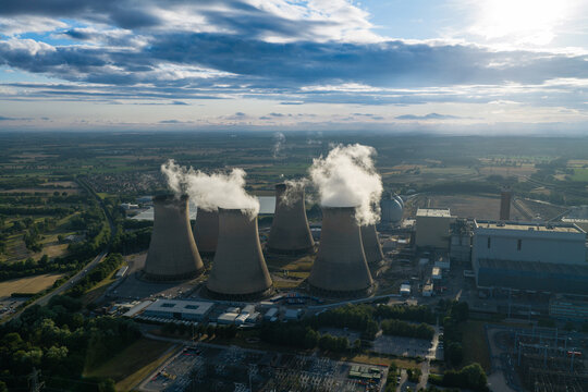 Aerial view of a nuclear power plant with cooling towers emitting steam, surrounded by rural landscape and fields under a dramatic cloudy sky in the United Kingdom.