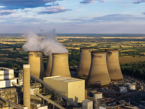 Aerial view of a power plant with large cooling towers emitting steam, surrounded by industrial infrastructure and expansive green fields in the United Kingdom.