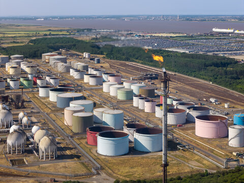Aerial view of an industrial oil refinery complex with numerous large storage tanks and a tall flare stack emitting a flame, set against a coastal landscape in the United Kingdom.