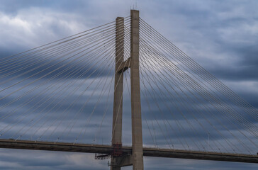 Fototapeta premium Wide view of a large cable-stayed bridge roadway supported by a central concrete tower and symmetrical steel cables under a dramatic, cloudy, blue-grey sky