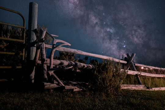 View of a bull elk antler resting on a rustic wooden ranch fence under the starry night sky and the Milky Way in Picabo, Idaho, United States.