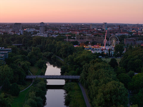 Hanover, Germany - 12 July 2026: Aerial view of Maschsee area with a fairground, lush trees, a river, and city skyline at sunset.